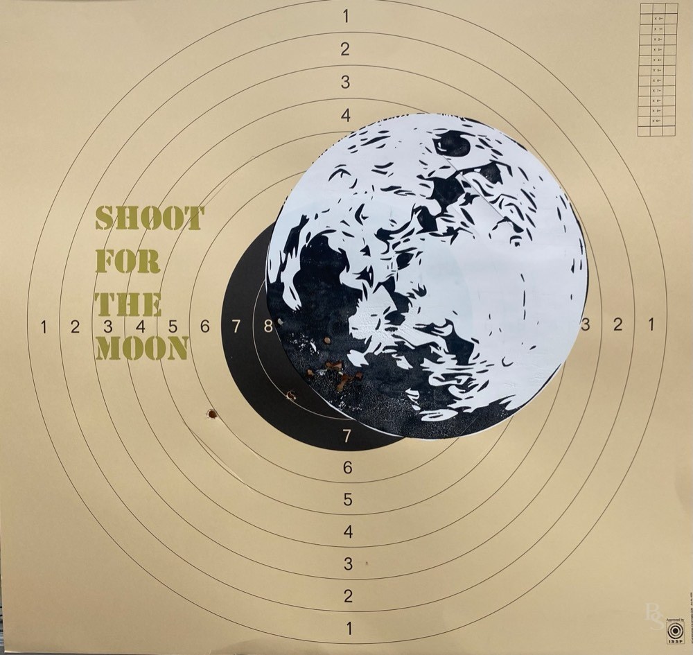 Siebdruck auf einer beschossenen Schießscheibe: Mond in Schwarz-Weiß, daneben der Schriftzug „Shoot for the Moon“